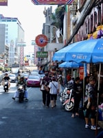 A busy city street filled with people walking and riding scooters. Neon signs with Thai writing hang from buildings, and numerous shops line the street. People are engaged in daily activities, creating a bustling atmosphere. A pink taxi can be seen amidst the crowd.