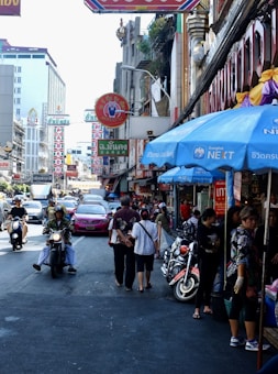 A busy city street filled with people walking and riding scooters. Neon signs with Thai writing hang from buildings, and numerous shops line the street. People are engaged in daily activities, creating a bustling atmosphere. A pink taxi can be seen amidst the crowd.