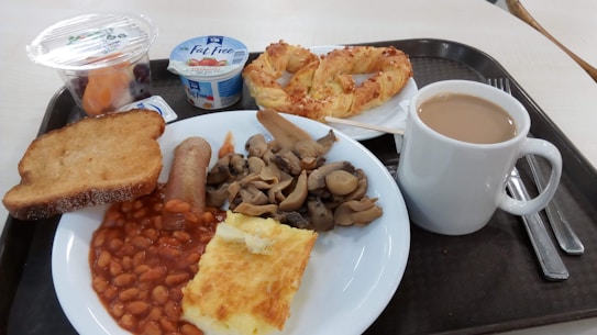 A breakfast tray contains a variety of food items including toast, baked beans, a sausage, mushrooms, scrambled eggs or omelette, a croissant-style pastry, a cup of coffee, yogurt, and a small container of fruit.