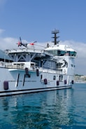 A large naval ship is berthed at a harbor with a helicopter parked on its deck. The ship's white and gray exterior contrasts with the calm blue waters. Several fenders hang along the side to protect it from the dock. The partly cloudy sky is visible in the background, and there are distant buildings along the waterfront.