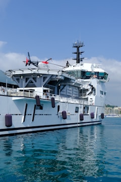 A large naval ship is berthed at a harbor with a helicopter parked on its deck. The ship's white and gray exterior contrasts with the calm blue waters. Several fenders hang along the side to protect it from the dock. The partly cloudy sky is visible in the background, and there are distant buildings along the waterfront.
