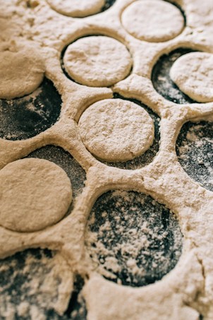 A close-up view of dough circles cut out from a rolled sheet of dough. The surface is dusted with flour, and the dough appears soft and ready for baking or further preparation.