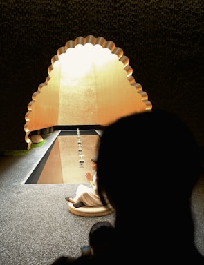 A person sits in a meditative pose on a round cushion, facing a pool of calm water. The scene is framed by an arched opening with scalloped edges, allowing soft, warm light to enter. The environment is serene and contemplative, suggesting a spiritual or reflective atmosphere.