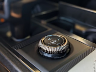 A close-up of a modern car's control knob located on the center console. The knob is marked with different vehicle mode options and is surrounded by textured edges for grip. The background includes part of the console with a sleek, dark finish.