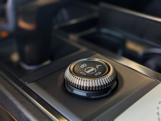 A close-up of a modern car's control knob located on the center console. The knob is marked with different vehicle mode options and is surrounded by textured edges for grip. The background includes part of the console with a sleek, dark finish.