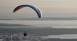 A paraglider is soaring in the sky above a cityscape with the sea and distant mountains in the background. The scene captures a sense of freedom and adventure.
