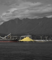 A large industrial site with a massive pile of yellow sulfur is located near a body of water. In the background, a series of towering mountains and a cloudy sky create a dramatic contrast. Structures, including a ship and cranes, dominate the industrial landscape.
