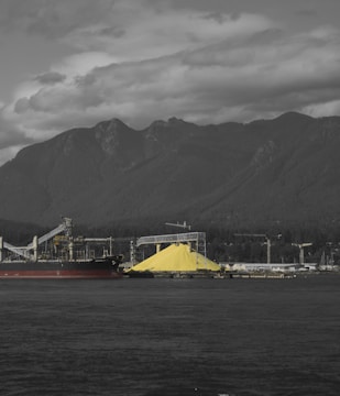 A large industrial site with a massive pile of yellow sulfur is located near a body of water. In the background, a series of towering mountains and a cloudy sky create a dramatic contrast. Structures, including a ship and cranes, dominate the industrial landscape.