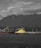 A large industrial site with a massive pile of yellow sulfur is located near a body of water. In the background, a series of towering mountains and a cloudy sky create a dramatic contrast. Structures, including a ship and cranes, dominate the industrial landscape.