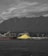 A large industrial site with a massive pile of yellow sulfur is located near a body of water. In the background, a series of towering mountains and a cloudy sky create a dramatic contrast. Structures, including a ship and cranes, dominate the industrial landscape.