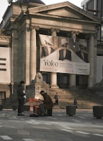 Two individuals are positioned in front of the Vancouver Art Gallery, with steps leading to the entrance and a large banner featuring black and white portraits. One person is playing a musical instrument while seated on an orange chair, while the other stands nearby. The architecture features classical columns and sculptures, and several pedestrians are visible in the background.