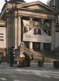Two individuals are positioned in front of the Vancouver Art Gallery, with steps leading to the entrance and a large banner featuring black and white portraits. One person is playing a musical instrument while seated on an orange chair, while the other stands nearby. The architecture features classical columns and sculptures, and several pedestrians are visible in the background.