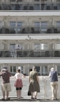 A multi-level cruise ship with passengers standing on different balconies. On the upper levels, people are visible behind glass railings. At the bottom level, five people stand with their backs to the camera, observing something off in the distance. The scene is well-lit with natural daylight.