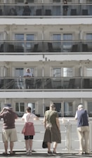 A multi-level cruise ship with passengers standing on different balconies. On the upper levels, people are visible behind glass railings. At the bottom level, five people stand with their backs to the camera, observing something off in the distance. The scene is well-lit with natural daylight.