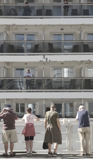 A multi-level cruise ship with passengers standing on different balconies. On the upper levels, people are visible behind glass railings. At the bottom level, five people stand with their backs to the camera, observing something off in the distance. The scene is well-lit with natural daylight.
