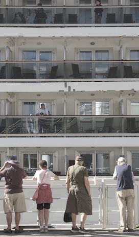 A multi-level cruise ship with passengers standing on different balconies. On the upper levels, people are visible behind glass railings. At the bottom level, five people stand with their backs to the camera, observing something off in the distance. The scene is well-lit with natural daylight.