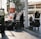 Two hotel staff members in uniform are handling luggage outside a building with a sign indicating a location in Canada Place. Several suitcases are stacked on trolleys. The background shows urban elements including trees, modern buildings, and a pedestrian walking in the distance.