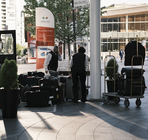 Two hotel staff members in uniform are handling luggage outside a building with a sign indicating a location in Canada Place. Several suitcases are stacked on trolleys. The background shows urban elements including trees, modern buildings, and a pedestrian walking in the distance.