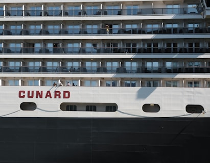 A cruise ship with multiple decks featuring rows of windows and balconies. The name 'Cunard' is prominently displayed in red on the side. There are a few people standing on one of the decks, with shadows cast across the ship's white and dark surfaces.