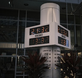 A large, white electronic clock displays digital time under the brand 'SEIKO.' The clock is mounted on a column surrounded by plants, with multiple city names indicating different time zones. The background shows a modern glass facade of a building with reflective windows.