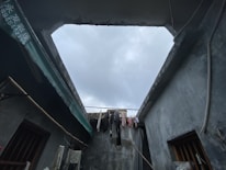 A partially open roof in a concrete structure reveals a cloudy sky above. Clothes are hanging on a line across the opening, indicating domestic activity. The setting appears to be somewhat rustic, with weathered walls and exposed wires. A green awning is visible on one side.