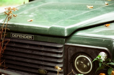 Close-up view of the front grille and headlight of a green Land Rover Defender with some dried plants and a few scattered leaves on the hood.