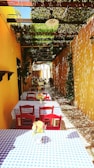 A charming outdoor cafe setting features a narrow walkway lined with tables covered in blue and white checkered cloths. Bright red chairs accompany each table, while flower arrangements in small vases add a touch of color. The space is shaded by a leafy canopy, casting intricate shadows on the ground and yellow walls, enhancing the rustic, Mediterranean ambiance.