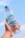A hand is holding a bottle of water against a clear blue sky. The bottle has a blue cap and a label that reads 'CRYSTALIN' with some graphics. It appears to be filled with transparent liquid.