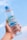 A hand is holding a bottle of water against a clear blue sky. The bottle has a blue cap and a label that reads 'CRYSTALIN' with some graphics. It appears to be filled with transparent liquid.