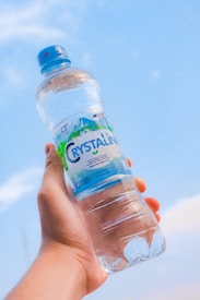 A hand is holding a bottle of water against a clear blue sky. The bottle has a blue cap and a label that reads 'CRYSTALIN' with some graphics. It appears to be filled with transparent liquid.