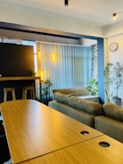 A cozy interior space featuring a comfortable sofa set against a large window with vertical blinds. Two wooden stools are positioned by a dark counter, illuminated by hanging pendant lights that add a warm glow. Indoor plants are interspersed throughout the room, contributing to a fresh and inviting atmosphere. A wall clock is visible on a light-colored wall, and exposed brick adds a rustic touch.