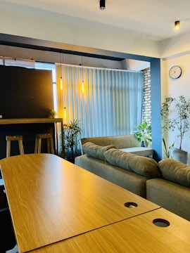 A cozy interior space featuring a comfortable sofa set against a large window with vertical blinds. Two wooden stools are positioned by a dark counter, illuminated by hanging pendant lights that add a warm glow. Indoor plants are interspersed throughout the room, contributing to a fresh and inviting atmosphere. A wall clock is visible on a light-colored wall, and exposed brick adds a rustic touch.
