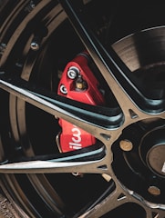 A close-up view of a car wheel with a black alloy rim and a prominent red brake caliper. The brake caliper has the word 'wil' visible on it, suggesting the brand Wilwood. The image focuses on the mechanical and metallic elements, with bolts and the brake disc partially visible.