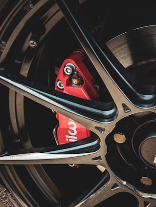 A close-up view of a car wheel with a black alloy rim and a prominent red brake caliper. The brake caliper has the word 'wil' visible on it, suggesting the brand Wilwood. The image focuses on the mechanical and metallic elements, with bolts and the brake disc partially visible.