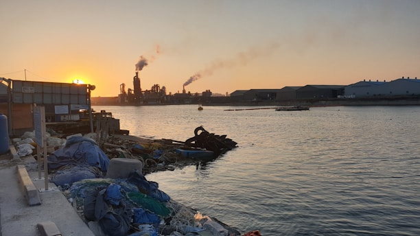 An industrial area by a body of water at sunset, with a factory emitting smoke into the air. A pile of debris and discarded items is visible in the foreground along the shoreline. The industrial buildings stretch across the horizon under the warm, orange glow of the setting sun.