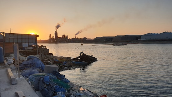 An industrial area by a body of water at sunset, with a factory emitting smoke into the air. A pile of debris and discarded items is visible in the foreground along the shoreline. The industrial buildings stretch across the horizon under the warm, orange glow of the setting sun.
