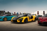 A lineup of three colorful sports cars parked on a concrete surface, with a blue sky background. The car in the center is a sleek yellow and black model, flanked by a vibrant blue car on the left with golden rims and a red car on the right. Banners and a tent are visible in the background, suggesting an outdoor event or exhibition.