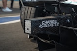 Close-up view of a racing car's rear section with carbon fiber details and a slogan 'CHANGE ACCELERATED' printed on it. The background includes blurred people and part of the racing track.