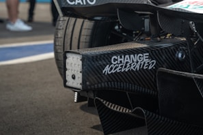 Close-up view of a racing car's rear section with carbon fiber details and a slogan 'CHANGE ACCELERATED' printed on it. The background includes blurred people and part of the racing track.