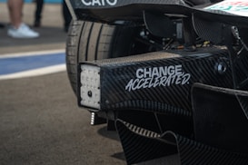 Close-up view of a racing car's rear section with carbon fiber details and a slogan 'CHANGE ACCELERATED' printed on it. The background includes blurred people and part of the racing track.