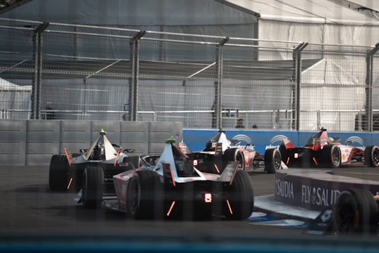 Several Formula E race cars are seen on a track with high barriers. The scene includes sleek, aerodynamic vehicles as they navigate a corner, with a metallic fence enclosing the racetrack. Banners and advertising signs are visible along the sides.
