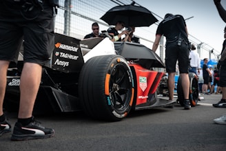 A sleek race car with prominent branding from various sponsors sits on a racetrack. Several individuals in racing attire surround the vehicle, with one person holding an umbrella. The scene appears to take place in a pit area with a fenced barrier in the background. The atmosphere is busy with activity, hinting at preparations for a racing event.