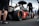A sleek race car with prominent branding from various sponsors sits on a racetrack. Several individuals in racing attire surround the vehicle, with one person holding an umbrella. The scene appears to take place in a pit area with a fenced barrier in the background. The atmosphere is busy with activity, hinting at preparations for a racing event.
