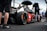 A sleek race car with prominent branding from various sponsors sits on a racetrack. Several individuals in racing attire surround the vehicle, with one person holding an umbrella. The scene appears to take place in a pit area with a fenced barrier in the background. The atmosphere is busy with activity, hinting at preparations for a racing event.