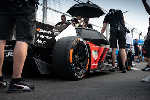 A sleek race car with prominent branding from various sponsors sits on a racetrack. Several individuals in racing attire surround the vehicle, with one person holding an umbrella. The scene appears to take place in a pit area with a fenced barrier in the background. The atmosphere is busy with activity, hinting at preparations for a racing event.
