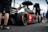 A sleek race car with prominent branding from various sponsors sits on a racetrack. Several individuals in racing attire surround the vehicle, with one person holding an umbrella. The scene appears to take place in a pit area with a fenced barrier in the background. The atmosphere is busy with activity, hinting at preparations for a racing event.