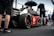 A sleek race car with prominent branding from various sponsors sits on a racetrack. Several individuals in racing attire surround the vehicle, with one person holding an umbrella. The scene appears to take place in a pit area with a fenced barrier in the background. The atmosphere is busy with activity, hinting at preparations for a racing event.