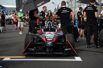 A race car is positioned on the track with several team members and personnel around it. The car is black and red with various sponsor logos. Some people are holding umbrellas providing shade, while others are dressed in uniforms. The background shows other people and elements of the racetrack.