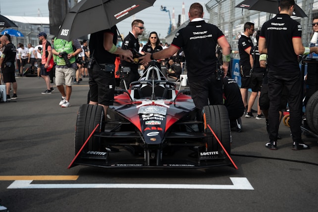 A race car is positioned on the track with several team members and personnel around it. The car is black and red with various sponsor logos. Some people are holding umbrellas providing shade, while others are dressed in uniforms. The background shows other people and elements of the racetrack.