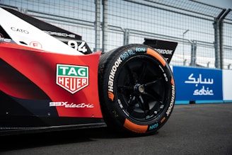 A close-up view of the rear wheel of a race car, prominently displaying the TAG Heuer logo and the word 'electric' on the side. The car has sleek, aerodynamic bodywork with visible branding. The wheel features Hankook tires with colorful markings. There is a wire fence and a blue advertisement board in the background.